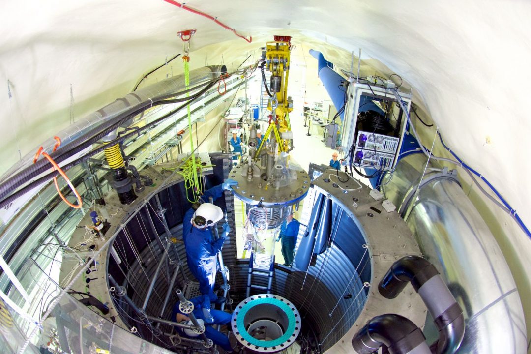 Inside a research facility, a person wearing a jumpsuit, gloves and hard hat works on an experiment from inside a cylindrical opening in the floor.