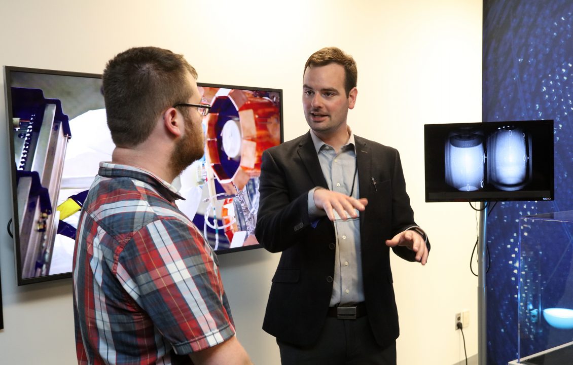 Two people speak while standing in front of a display with images of experiments.