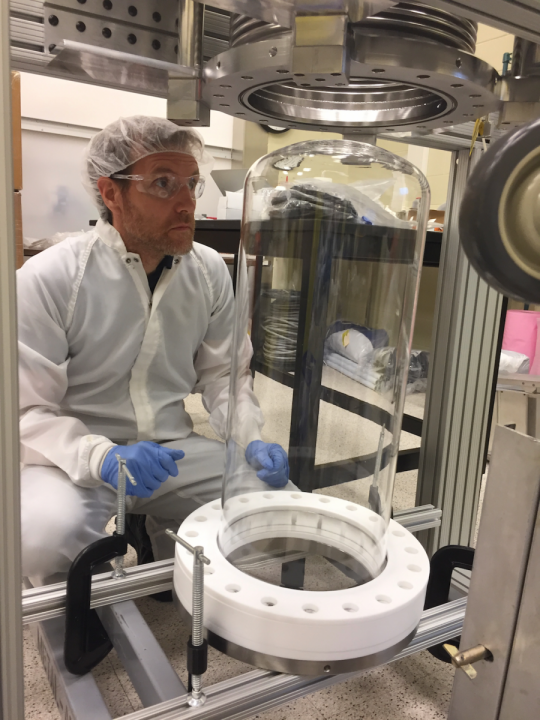 Inside a research facility, a person wearing a hairnet, safety goggles, a lab coat and gloves looks up into a cylindrical cavity. Below the cavity is a glass cylinder sitting on a metal frame.