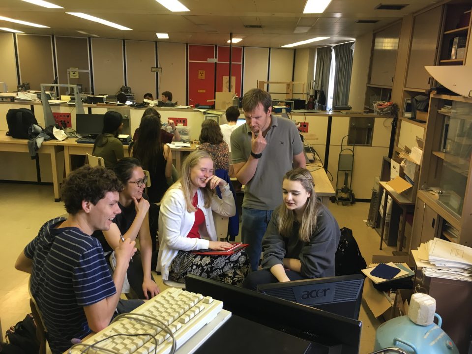 Five people speak to each other in a group, while sitting inside an office.