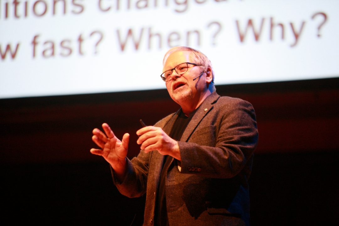 John Smol speaks into a microphone on a stage. A large presentation slide is visible in the background.