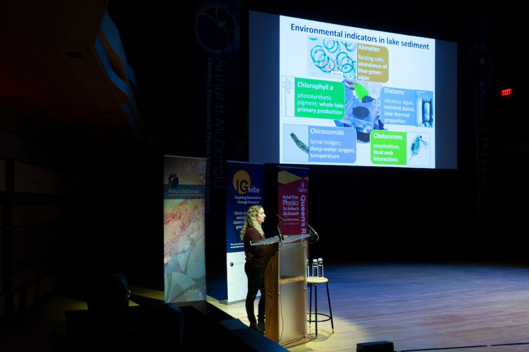 Liz Favot speaks into a microphone while standing at a podium. A presentation projected above is titled 