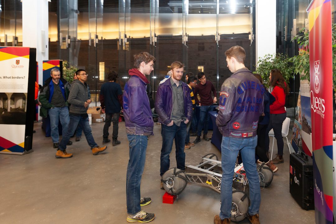 Three people stand around a robotic vehicle. In the background, there are various signs and people walking around.