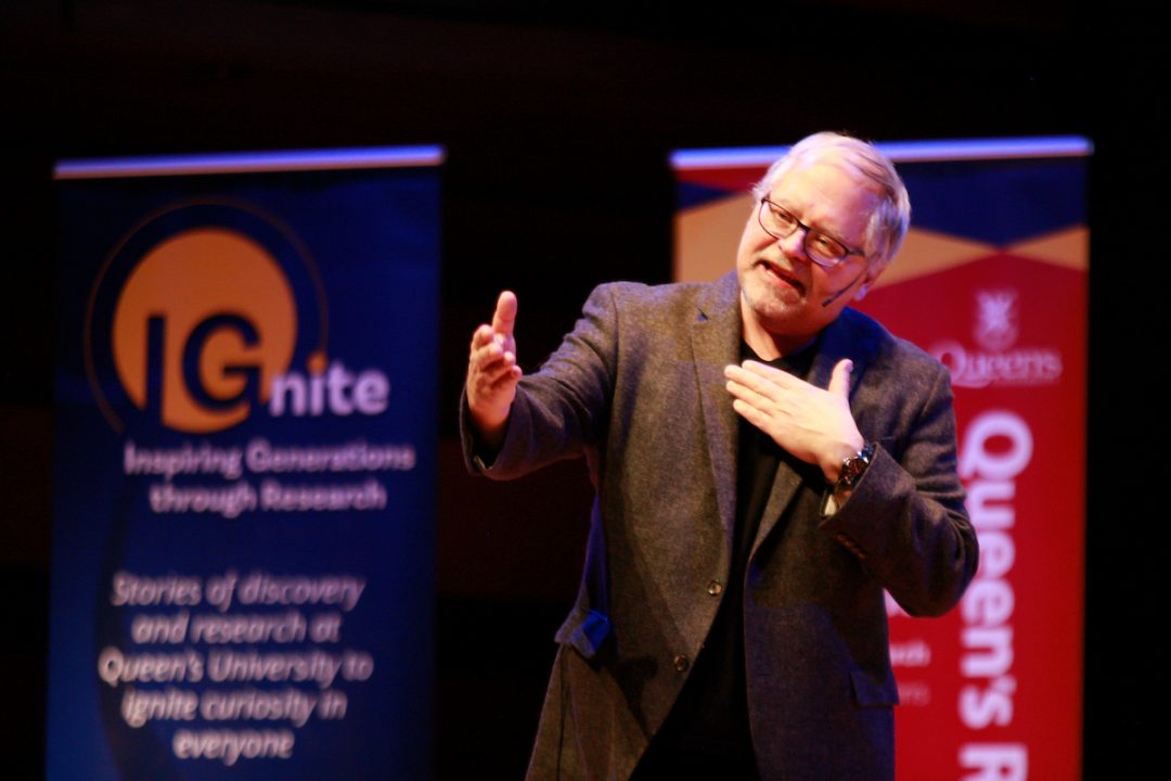 John Smol speaks into a microphone on a stage. Signs reading 