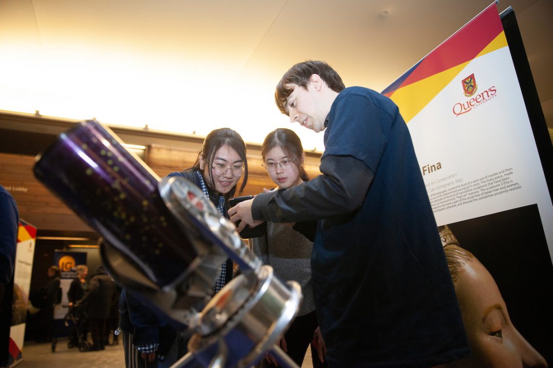 Two people look at an object in Connor's hand. In the foreground, a telescope is visible.