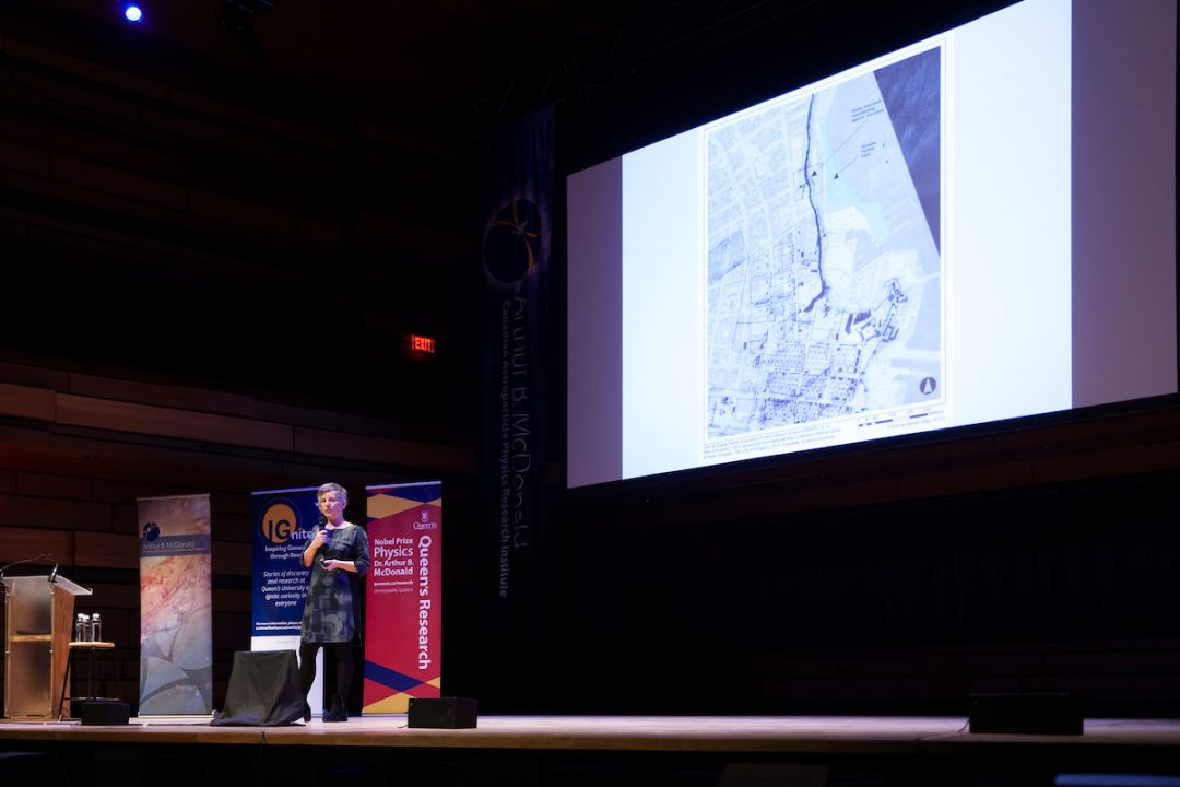 Laura Murray speaks into a microphone while standing on a stage. Several signs are visible in the background, reading IGnite and Queen's Research. Above, a large presentation slide displays a map