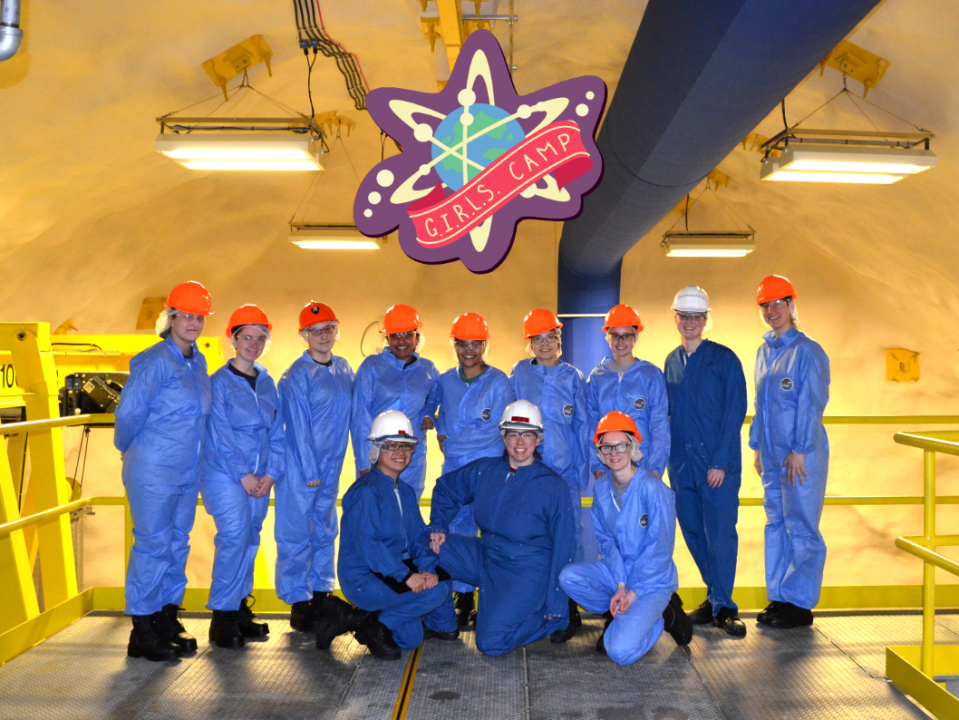 A group of people wearing personal protective equipment (hard hats, safety goggles and jumpsuits) pose together in a research facility. Text reads 