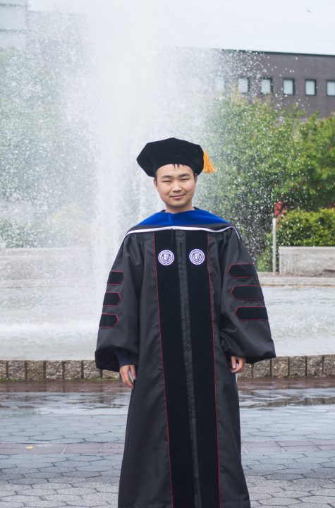 Ningqiang Song is pictured in front of a water fountain, wearing a graduation gown and cap