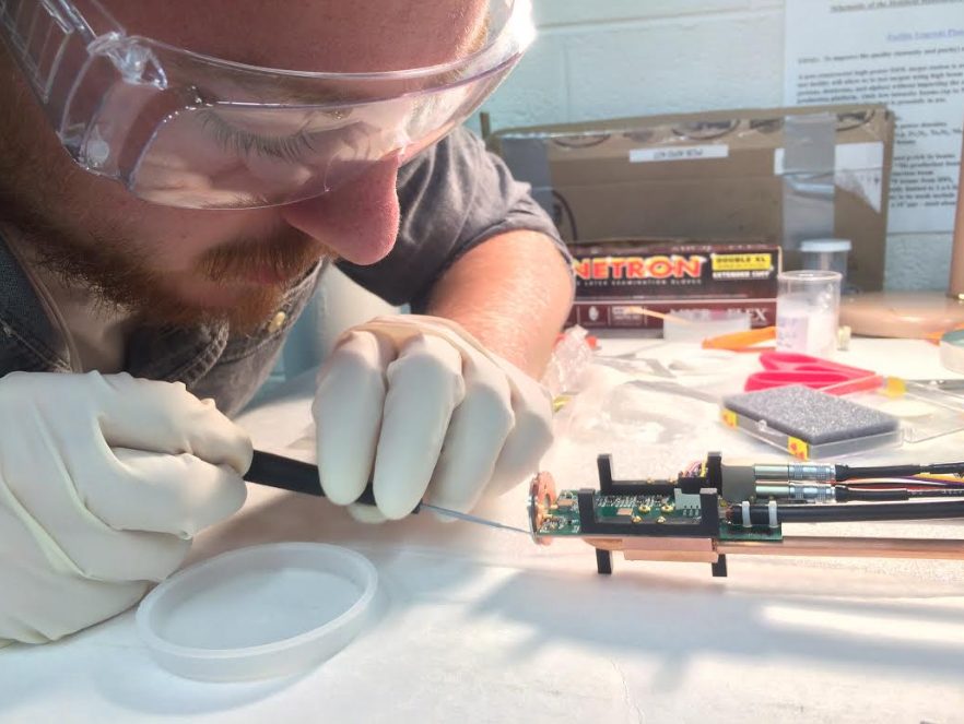 Matt is shown wearing lab goggles and gloves, using a tool to do fine-scale work on a small device