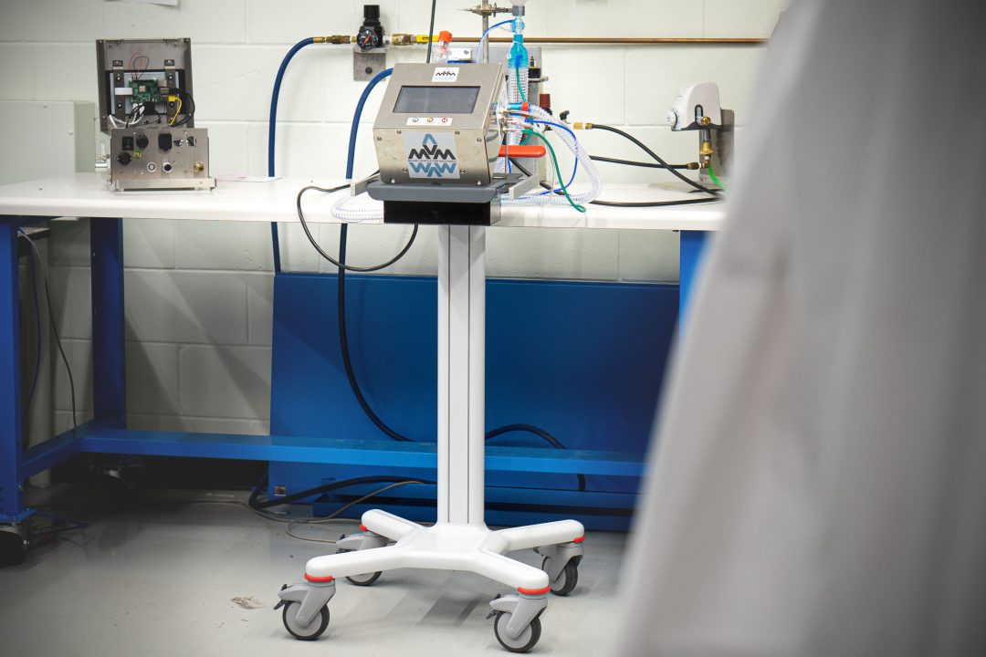 A mechanical ventilator (which looks like a metal box with a screen and buttons on it) sits on a rolling table, hooked up to many wires.