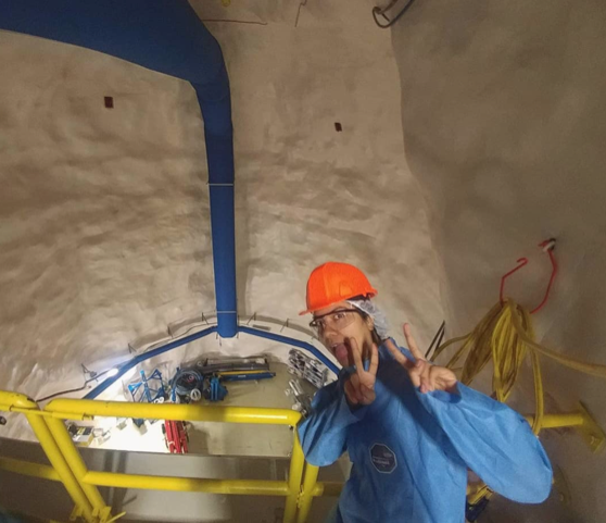 A person wearing personal protective equipment (hard hat and jumpsuit) poses inside a research laboratory and holds up peace signs