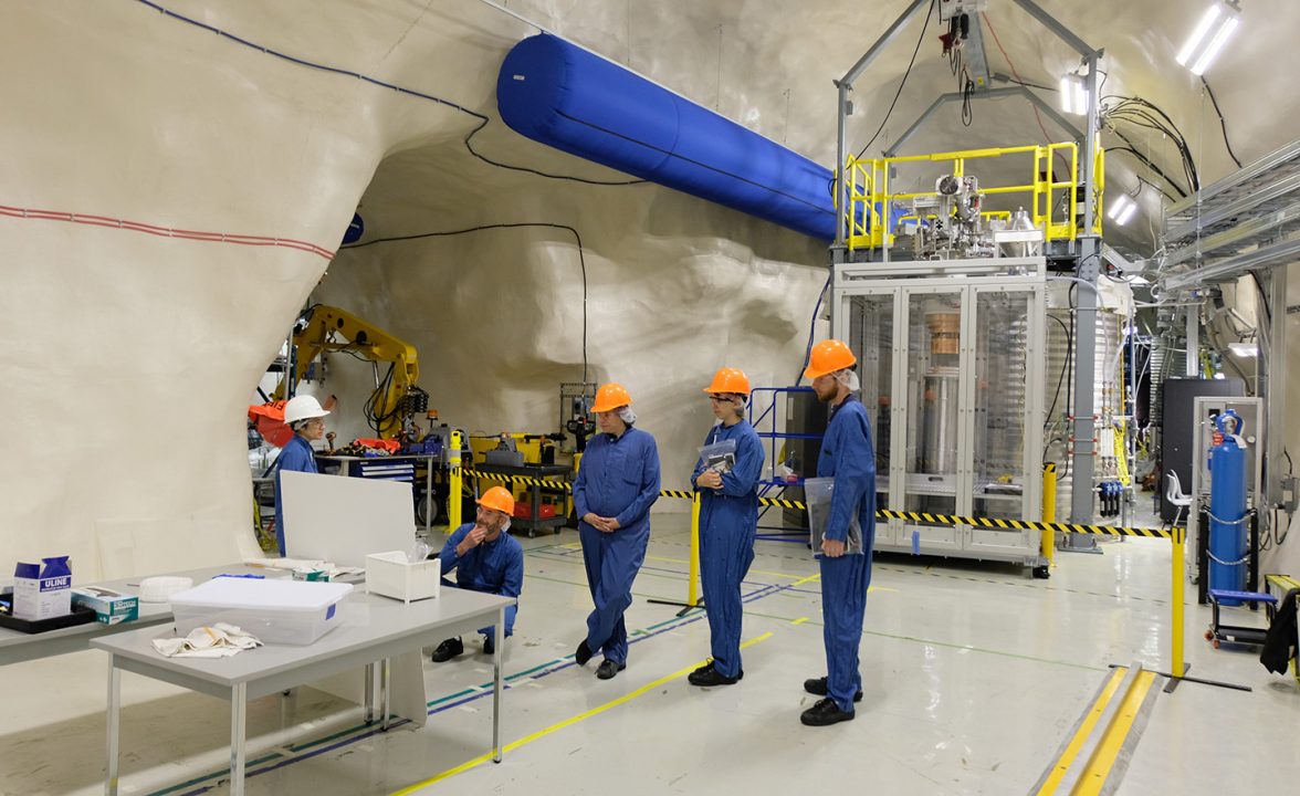 People wearing personal protective equipment (hard hats and jumpsuit) look at a computer inside a research facility containing many metal parts, equipment, tables and a gas cylinder