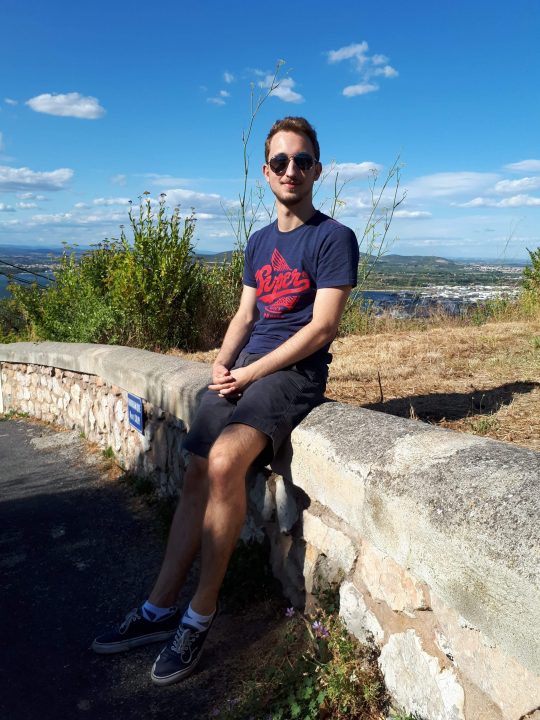 Lucas Feneaux sitting on a stone wall with landscape and blue skies in the background.