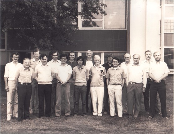 A black and white photo depicts people outside a building, standing in two rows while posing together for the photo