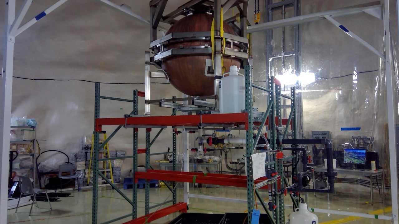 The SNOGLOBE copper sphere on an elevated platform. A copper sphere is in the upper centre of the image, with scaffolding underneath. The background of the image is a cleanroom, with a bright light and various equipment on tables.