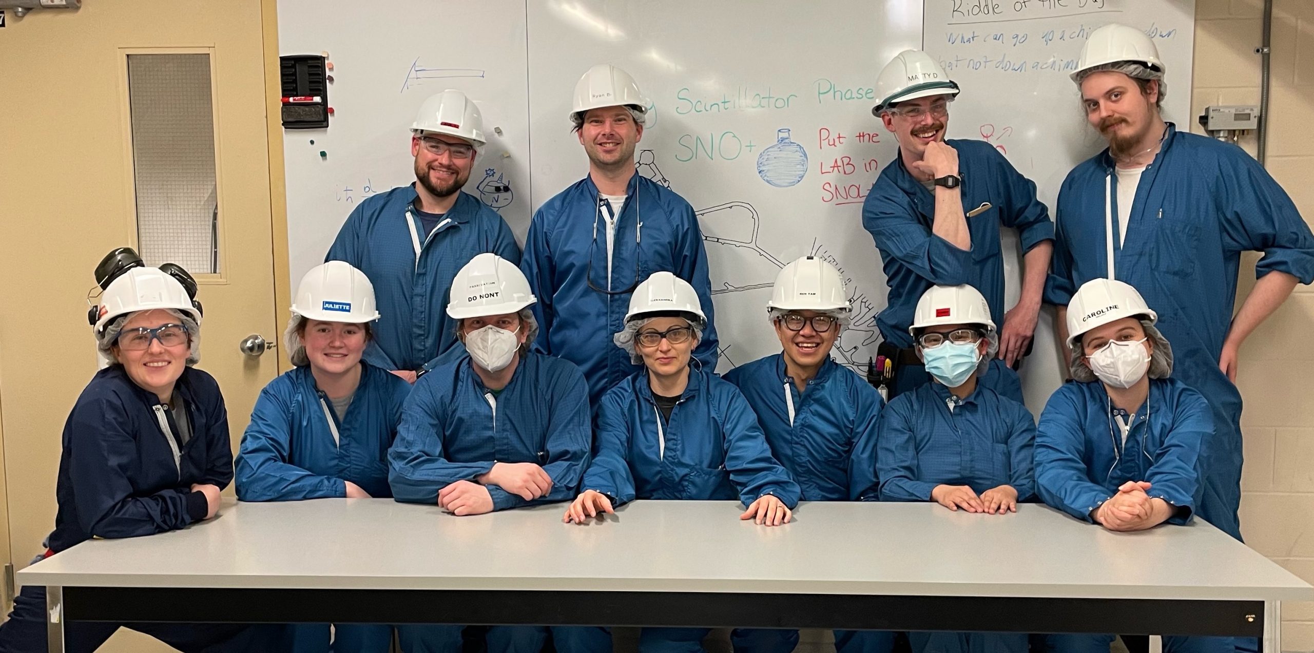 The SNO+ underground team smiles and sits (front row) and stands (back row) behind a large table and in front of a large whiteboard. Everyone is wearing blue or black coveralls and white hardhats.