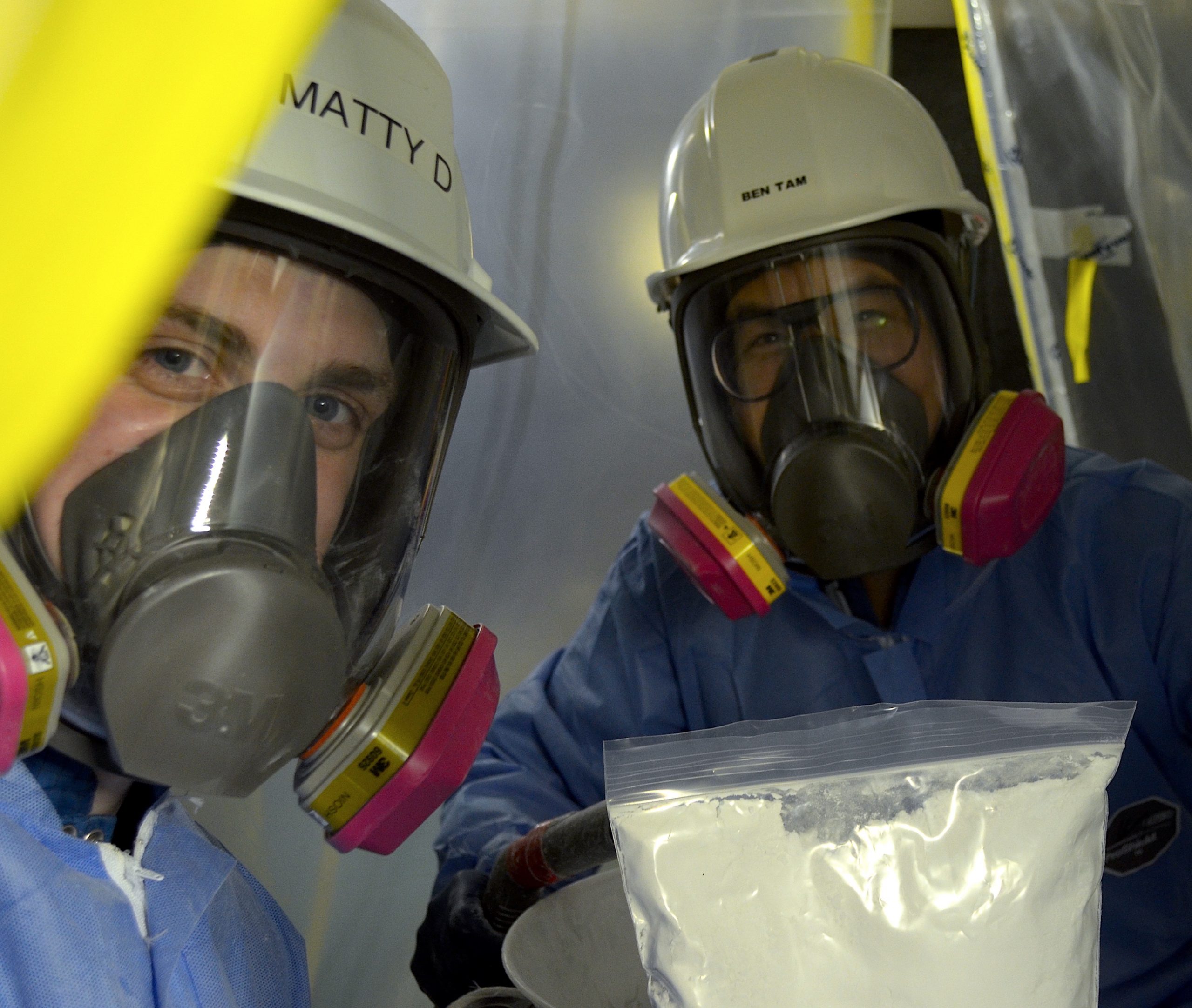 Two people in full personal protective equipment (jumpsuits, gas masks with face shields, hard hats, gloves) are in a compact space. They are smiling while one person holds a large plastic baggie of white powder.