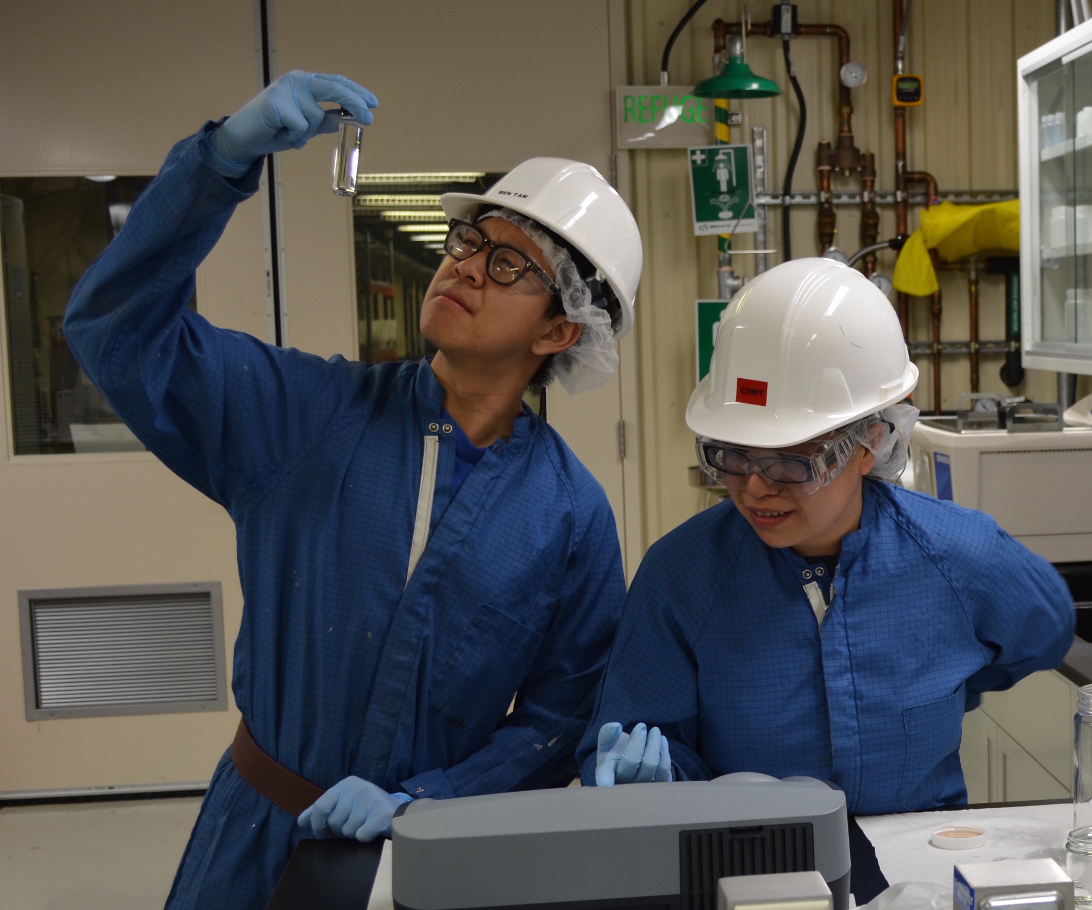 Two people wearing blue coveralls, white hardhats, latex gloves, safety glasses, and hairnets are working in a room with scientific equipment. One person is holding a tube of clear liquid up to the light and the other is pointing to a monitor.