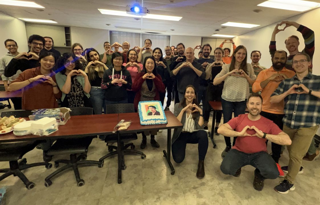 A large group of people from the Queen's astroparticle physics group pose smile for the camera and create heart shapes with their hands, while celebrating International Women's Day
