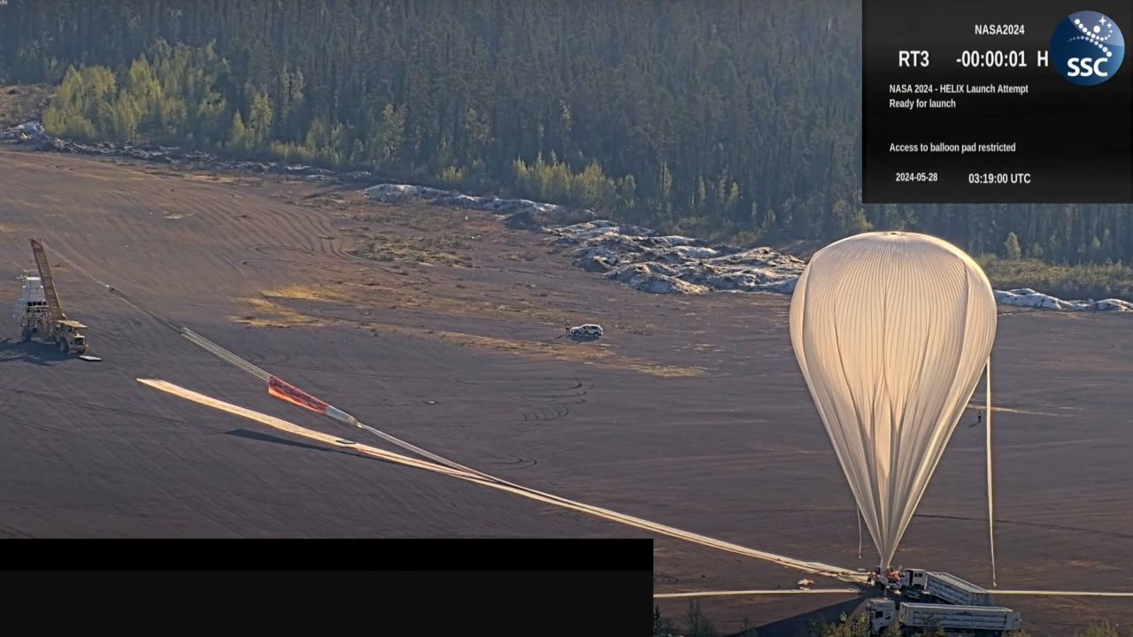 A screenshot from the youtube livestream of the HELIX experiment launch shows a large white balloon on a launchpad. On the other end of the launch area, a truck with a crane holds the thether for the payload upright.