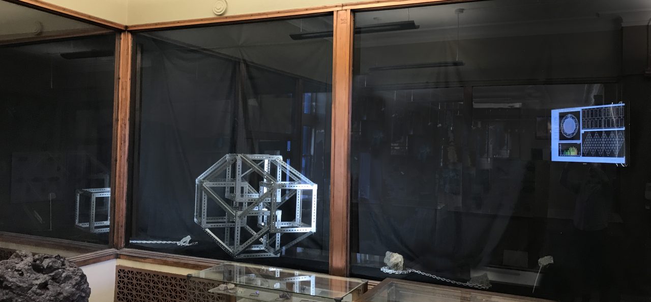 A display case with multiple sculptures. Small rock arrangements are in the bottom and a large polyhedral shape made of metal strapping sits in the center. A video screen plays a recording of the SNO+ experiment readout.