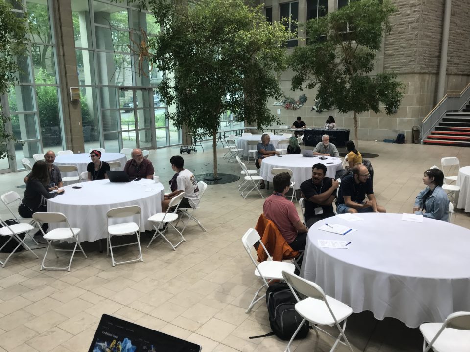 Small groups of physicists and students sit around round tables in a spacious atrium at Queen's University for the 2023 McDonald Institute National Meeting HQP Workshop