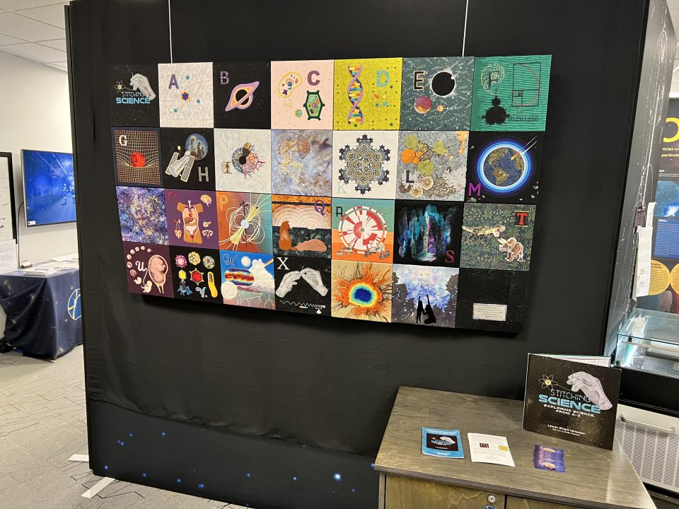 photo of an art installation featuring 28 individual 10 inch by 1 inch artworks of hand stitched/embroidered science topics.