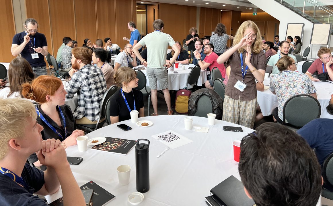A large room with multiple round tables. Groups of physics students sit around each table and one person from each table is standing, performing charades.