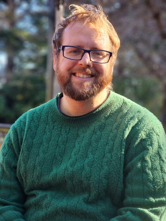 Caleb Miller smiling wearing a green sweater in an outdoor setting.